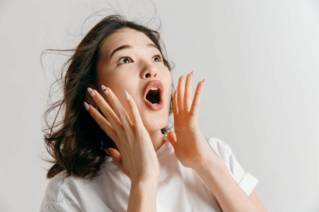 Wow. Beautiful female half-length front portrait isolated on gray studio backgroud. Young emotional surprised asian woman standing with open mouth. Human emotions, facial expression concept.